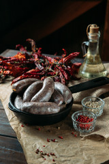 Homemade juicy pork sausages in a black frying pan, spices, pickled tomatoes and dried peppers on a craft paper, on wooden rustic background.