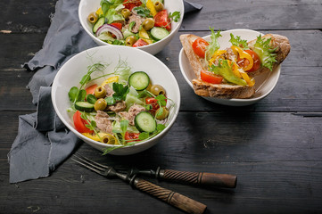 Close-up salads with vegetables and micro-greens in white bowls, canapes with pate and sweet pepper on a dark wooden table.