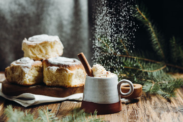 A hot cocoa with cinnamon stick and fresh Christmas buns with powder and over festive table, sugar powder is sprinkled on the background lke a snow. Winter holidays.