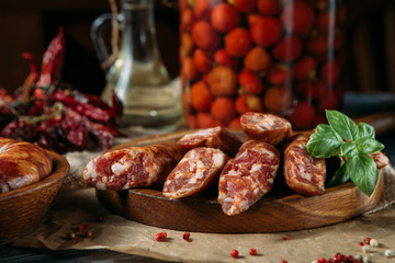 Horizontal photo of homemade raw chopped sausages in a wooden bowl, dried peppers and pickled tomatoes on the wooden table.