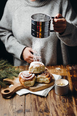Woman in grey sweater sprinkling sugar powder onto cinnamon buns, lying on the wooden board. Christmas mood with fur-tree branches.