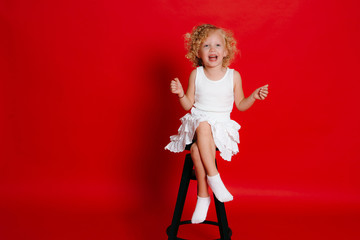 Amuzing funny little blonde girl in white dress sitting on chair isolated on red