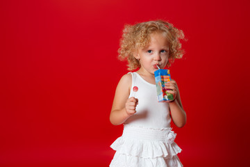 Cute curly girl in white dress drinking juice holding lollipop isolated on red background