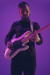 Young and joyful caucasian musician playing guitar on gradient purple studio background in neon light. Concept of music, hobby, festival. Colorful portrait of modern artist. Attented and inspired.