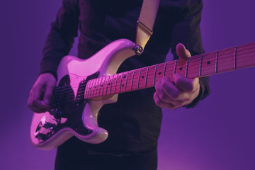 Fototapeta na wymiar Young and joyful caucasian musician playing guitar on gradient purple studio background in neon light. Concept of music, hobby, festival. Colorful portrait of modern artist. Attented and inspired.