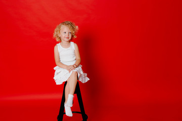 Modest little blonde girl in white dress sitting on chair isolated on red