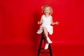 Amuzing funny little blonde girl in white dress sitting on chair isolated on red