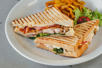Plate with two chicken and bacon club sandwiches with fries and vegetables on a white plate on a concrete background.