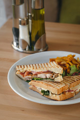 Close-up of chicken and bacon club sandwich with fries and vegetables on a white plate on a wooden background.