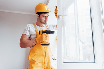 Handyman in yellow uniform installs new window by using automatic screwdriver. House renovation...