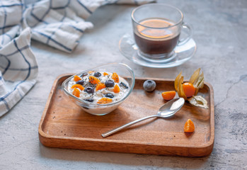 Breakfast with coffee and yogurt in a glass bowl with blueberries and physalis on a wooden brown tray on a concrete gray-blue background with a towel in a thin blue check.