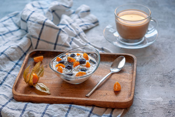 Breakfast with coffee and yogurt in a glass bowl with blueberries and physalis on a wooden brown tray on a concrete gray-blue background with a towel in a thin blue check.
