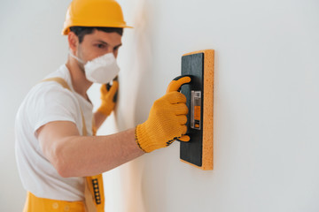 Handyman in yellow uniform and protective mask polishing wall indoors. House renovation conception