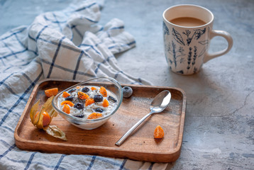 Breakfast yogurt in a glass bowl with blueberries and physalis on a wooden brown tray on a concrete gray-blue background with a towel in a thin blue check.