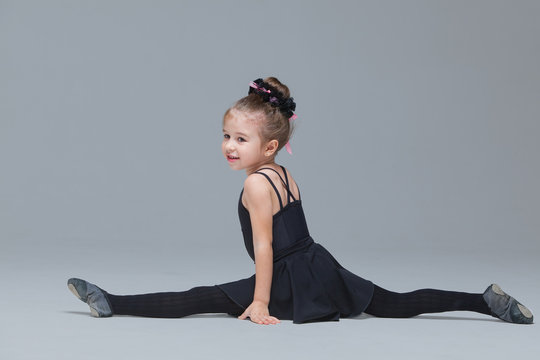 Beautiful little gymnast in black sportswear shows exercises on grey background. Flexible girl doing the splits to become ballet dancer