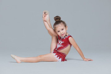 Flexible little girl in beautiful red dress is stretching doing gymnastic exercises on grey...
