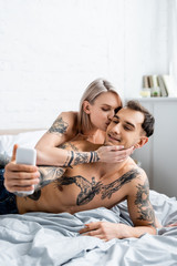 Beautiful girl kissing shirtless tattooed boyfriend taking selfie with smartphone on bed