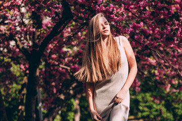 Closeup portrait of a lovely girl in a park with blooming Japanese sakura trees. Romantic young blonde in a dress posing on a background of spring flowering trees. Cherry blossoms.