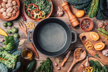 Various organic vegetables ingredients and empty iron cooking pot, wooden bowls, spoons on wooden background. Top view, copy space. Organic vegetables ingredients for vegan cooking. Clean eating food