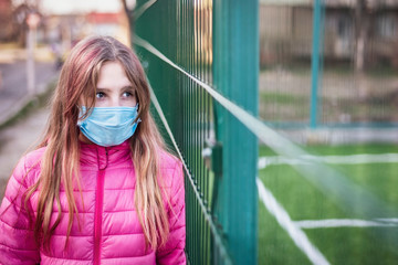 A sad teenage girl in a medical mask stands near a football field fenced in with a net. Now there is a quarantine and the girl is sad because she never saw the football field empty