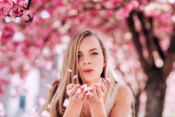 Closeup portrait of a lovely girl in a park with blooming japanese sakura trees. Romantic young blonde in a dress posing on a background of spring flowering trees. Flying cherry petals.