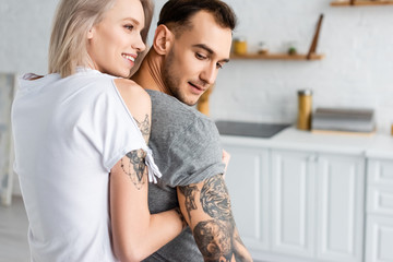 Back view of smiling woman embracing tattooed boyfriend in kitchen