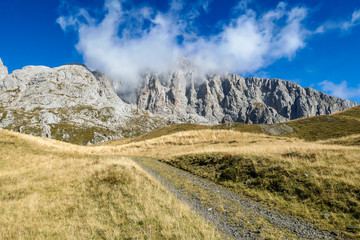 Fototapeta na wymiar A pathway leading to high peaks in Italian Alps. Sharp slopes on both sides of the valley. Hard to reach mountain peaks. There are many mountain ranges in the back. Serenity and peace. Autumn vibes