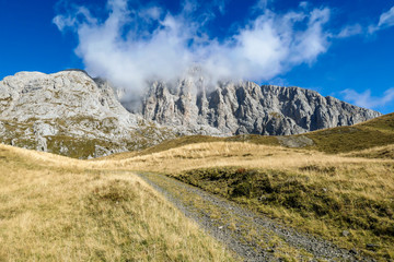 Fototapeta na wymiar A pathway leading to high peaks in Italian Alps. Sharp slopes on both sides of the valley. Hard to reach mountain peaks. There are many mountain ranges in the back. Serenity and peace. Autumn vibes