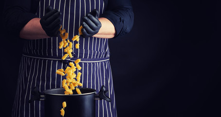 chef in black latex gloves, striped apron pours raw fusilli pasta into a metal pan