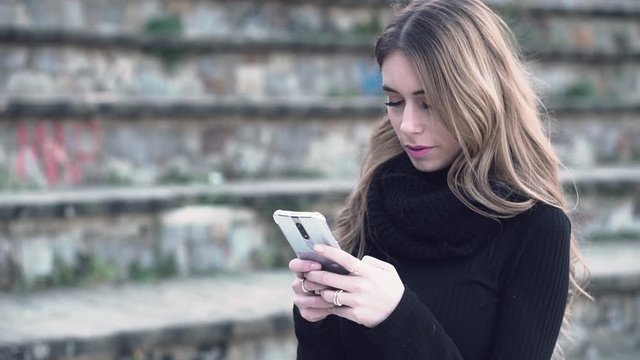 Young blonde caucasian woman standing outdoors texting and reading messages on her mobile smart phone waiting for her date