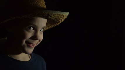 A boy is standing in the dark. On the head is a straw cowboy hat. Side view. Glance looks at the light source. Close-up.