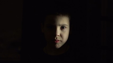 The boy's face is illuminated. Open eyes. Look straight. Restrained emotions. Dark room. Horror....