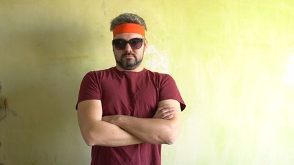 A man with a beard and dark glasses. Standing at the wall. Dark t-shirt. The orange tape on the head. Important kind. A confident pose. The movement of the hands. Freak.