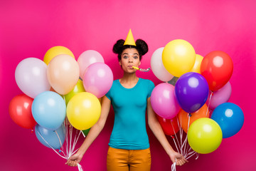 Photo of pretty funny dark skin lady hold many colorful air balloons blow noisemaker birthday surprise wear paper cone cap blue t-shirt yellow pants isolated bright magenta color background