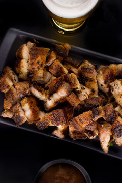 Grilled pork belly in a black platter with barbecue salce and beer in black background seen from above