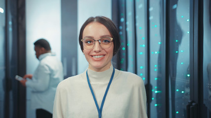 Young attractive woman IT engineer with tablet smiling at camera standing near rack server cages cooperating in data center security storage.