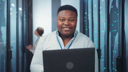 Portrait of nice cheerful african administrator with notebook enjoying modern job working in digital server room at data server farm. Database. Information technology.