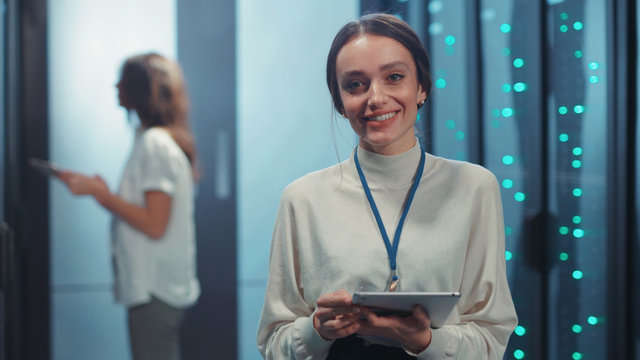 Joyful pretty young woman IT technician holding tablet staying at modular data center. Portrait of smiling female specialist coworking at rack server room.