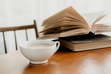 a white Cup of coffee with milk stands on a wooden table with an open book in the background. light from the window. the concept of reading books over a Cup of morning coffee