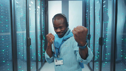 Excited black IT administrator dancing in server rack corridor happy with finished work having fun time at data center.