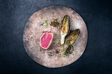 Fried dry aged beef fillet medallion steak natural with artichokes and herbs as top view on a rustic modern design plate with copy space