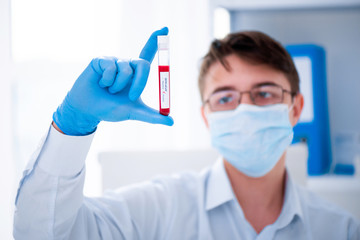 Test tube in male hand close up, doctor in medical mask holding vial with red liquid. Concept blood...