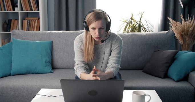 Concentrated mature woman in casual clothing sitting with headset on couch and having video conference on laptop. Beautiful lady doing remote work at home.