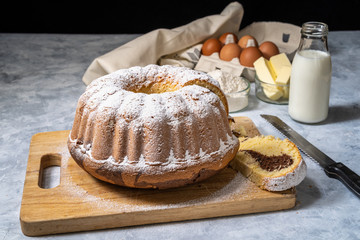 Fresh bundt cake decorated with icing sugar on a wooden cutting board with ingredients in a background. Front view of a delicious homemade marble cake baked in a mold.