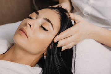 Close up photo of a brunette caucasian woman lying with closed eyes while having a head massage at the spa salon