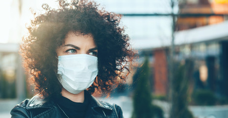 Curly haired caucasian woman wearing an anti flu mask while posing outside on front of a building