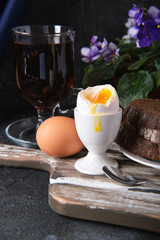 Two boiled eggs on a cutting board spoon, pieces of rye bread on a plate, a glass of tea and a flower in a pot on a dark table. Breakfast.