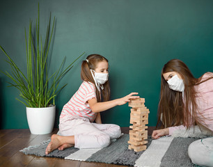 Two sisters in medical mask playing playing a blocks wood tower game (jenga) at home during covid quarantine. Stay at home activity for kids concept.