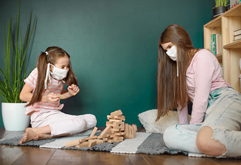 Two sisters in medical mask playing playing a blocks wood tower game (jenga) at home during covid quarantine. Stay at home activity for kids concept.