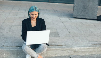Caucasian lady with blue hair posing outside with a computer while sitting and working